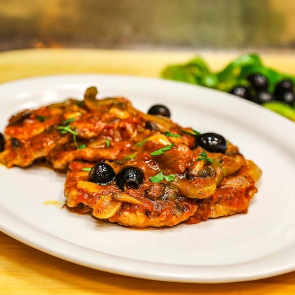 Plated pasta topped with tomato sauce, black olives, mushrooms, and fresh herbs, with greens in the background.